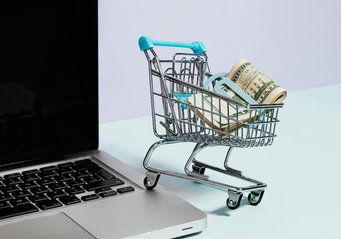 A shopping cart with money, on the edge of a laptop