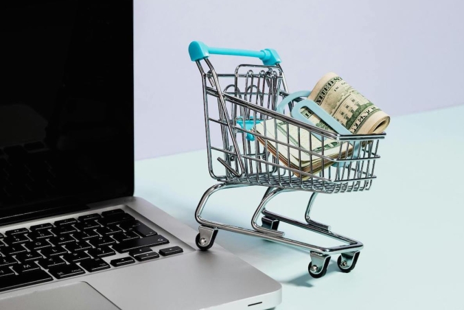 A shopping cart with money, on the edge of a laptop
